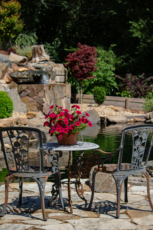 Two chairs on a flagstone patio overlooking a pond with a waterfall in the background.